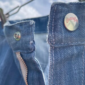 Pu&ograve; includere: Primo piano di jeans di denim blu, che mostra i dettagli del bottone, della cerniera e delle cuciture. Il bottone ha la scritta "Wrangler" in lettere rosse. I jeans sono appesi a una gruccia di metallo.