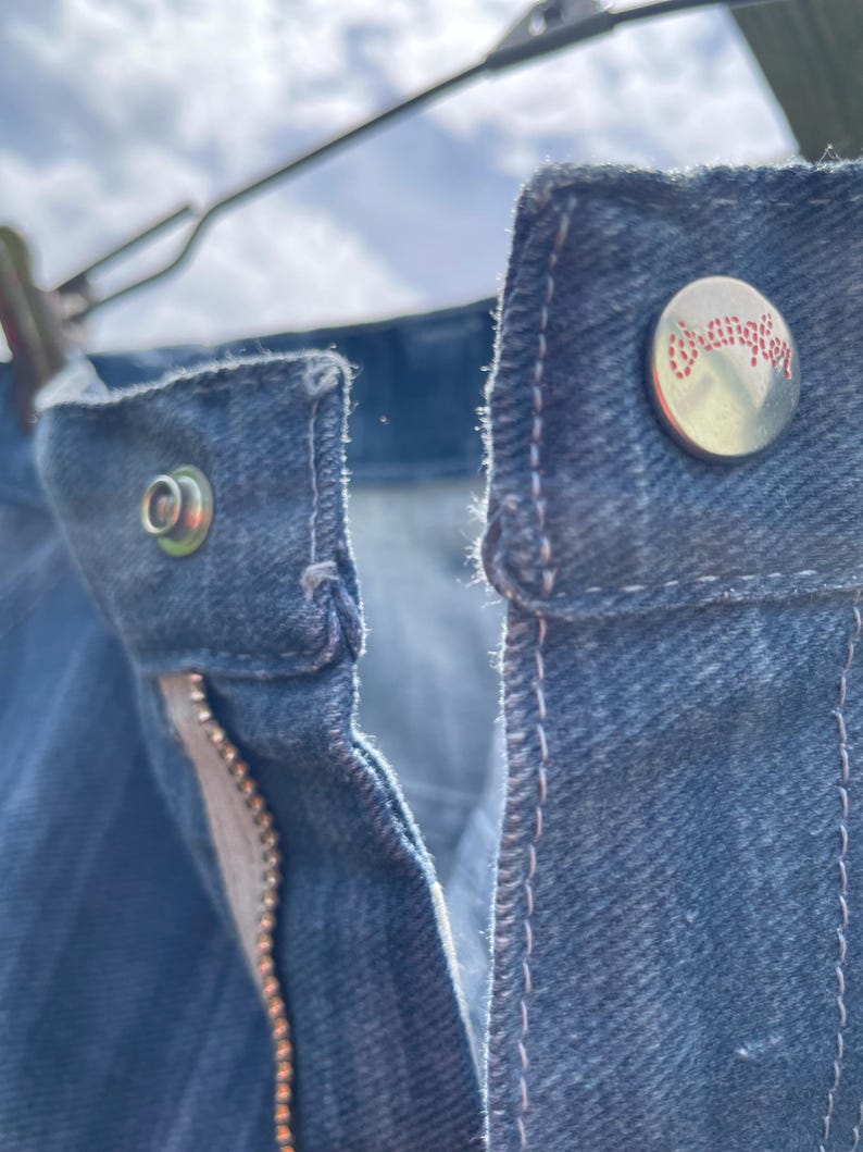 Pu&ograve; includere: Primo piano di jeans di denim blu, che mostra i dettagli del bottone, della cerniera e delle cuciture. Il bottone ha la scritta "Wrangler" in lettere rosse. I jeans sono appesi a una gruccia di metallo.