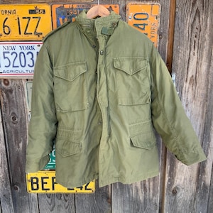 May include: Olive green military-style jacket with multiple front pockets and a button-up closure. The jacket has a collared neckline and long sleeves. The jacket is hanging on a wooden hanger.