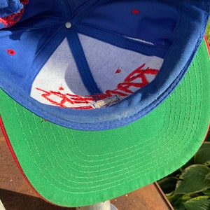 May include: A blue and green baseball cap with red stitching. The cap has a white mesh lining and red embroidery on the front.