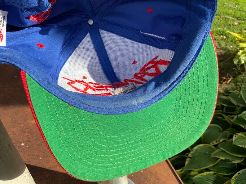 May include: A blue and green baseball cap with red stitching. The cap has a white mesh lining and red embroidery on the front.