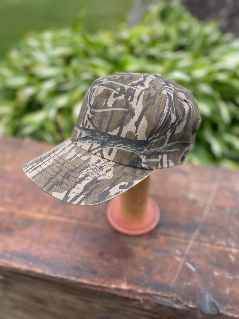 Op de afbeelding: Een baseballpet met een bruin en beige camouflagepatroon met een boomschorsontwerp. De pet heeft een gestructureerde kroon en een gebogen klep. Een klein logo met de tekst Mossy Oak is gedrukt op de linkerkant van de klep bij de kroon.