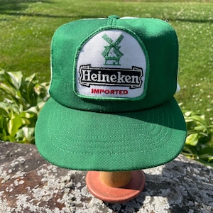 May include: A green and white trucker hat featuring a central patch with a green windmill icon above the black text Heineken and the red word IMPORTED. The cap has a solid green front panel and a white mesh back panel with a curved green brim.