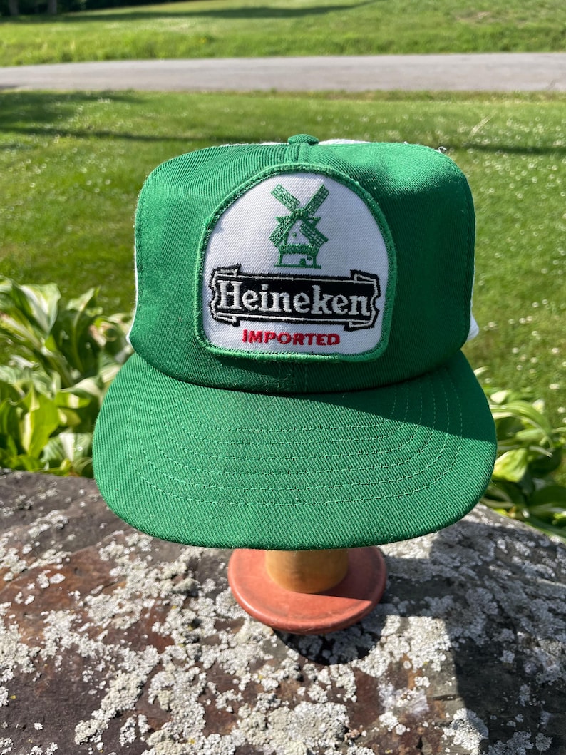 May include: A green and white trucker hat featuring a central patch with a green windmill icon above the black text Heineken and the red word IMPORTED. The cap has a solid green front panel and a white mesh back panel with a curved green brim.