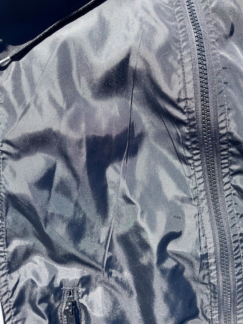 Puede incluir: Primer plano de una chaqueta negra con una textura brillante y ligeramente arrugada. La imagen muestra una cremallera de cuerpo entero en el lado derecho y una cremallera m&aacute;s peque&ntilde;a en la parte inferior. La chaqueta parece estar hecha de un material ligero.