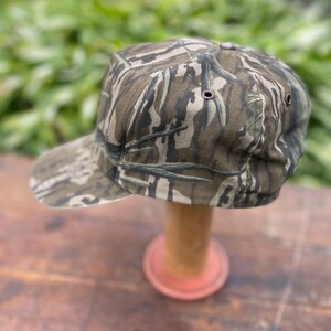 Op de afbeelding: Een baseballpet met een camouflagepatroon in donkergroen, bruin en zwart. De pet heeft een gestructureerde kroon, een gebogen klep en kleine metalen oogjes aan de zijkanten voor ventilatie. De stof vertoont een gedetailleerde boomschorsstructuur.