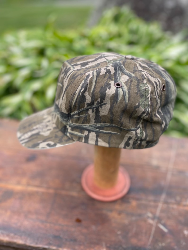 Op de afbeelding: Een baseballpet met een camouflagepatroon in donkergroen, bruin en zwart. De pet heeft een gestructureerde kroon, een gebogen klep en kleine metalen oogjes aan de zijkanten voor ventilatie. De stof vertoont een gedetailleerde boomschorsstructuur.