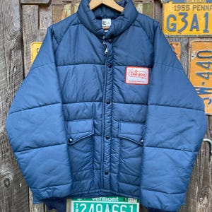 May include: A vintage navy blue quilted jacket with a high collar and button closure. Features two front pockets and a Dairylea patch. The jacket is hanging on a wooden hanger. The background includes vintage license plates.