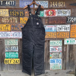 May include: Black Carhartt overalls with adjustable straps and multiple pockets. The workwear garment is hanging on a wooden hanger against a backdrop of vintage license plates. The overalls have brass-colored rivets.