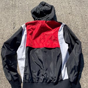 Puede incluir: Chaqueta cortavientos con capucha negra, roja y blanca. La chaqueta tiene capucha, mangas y dobladillo negros. Un panel rojo est&aacute; centrado en la parte superior de la espalda, con paneles blancos a cada lado. La chaqueta est&aacute; hecha de un material ligero, posiblemente nailon.