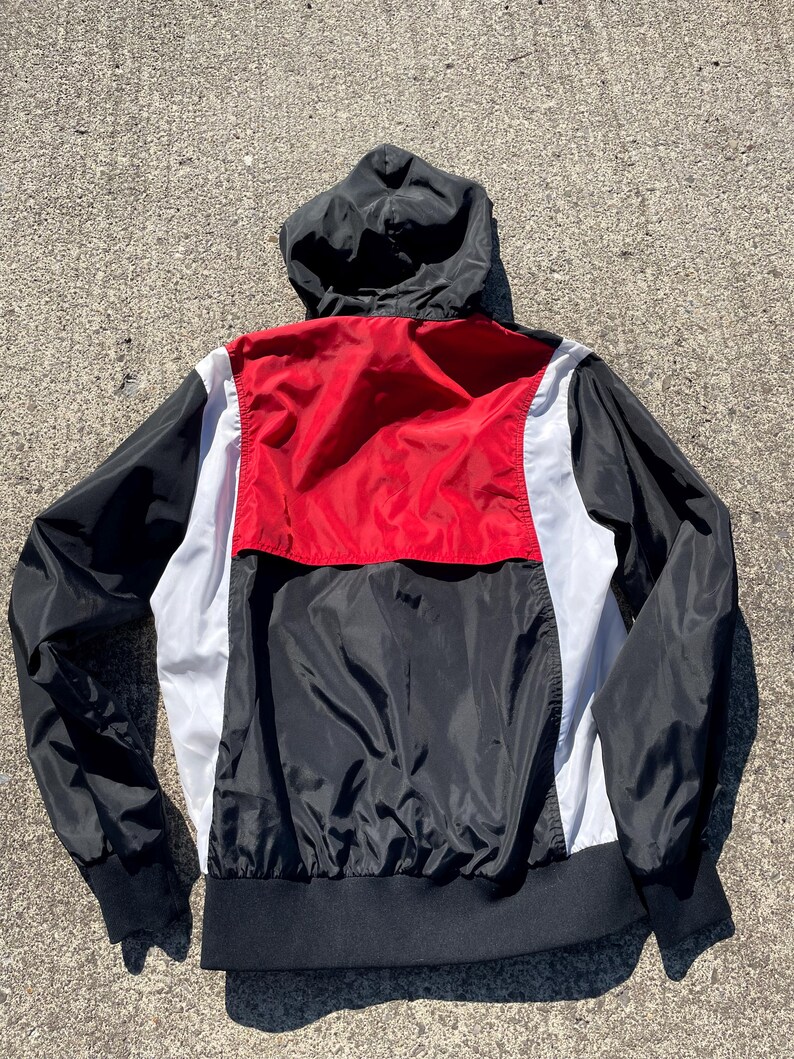 Puede incluir: Chaqueta cortavientos con capucha negra, roja y blanca. La chaqueta tiene capucha, mangas y dobladillo negros. Un panel rojo est&aacute; centrado en la parte superior de la espalda, con paneles blancos a cada lado. La chaqueta est&aacute; hecha de un material ligero, posiblemente nailon.