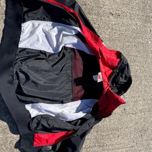 Puede incluir: Una chaqueta cortavientos Nike con un dise&ntilde;o de bloques de color en negro, blanco y rojo. La prenda cuenta con un cuello alto, cierre de cremallera frontal y un forro interior blanco. Una peque&ntilde;a etiqueta de la marca Nike es visible cerca del cuello.