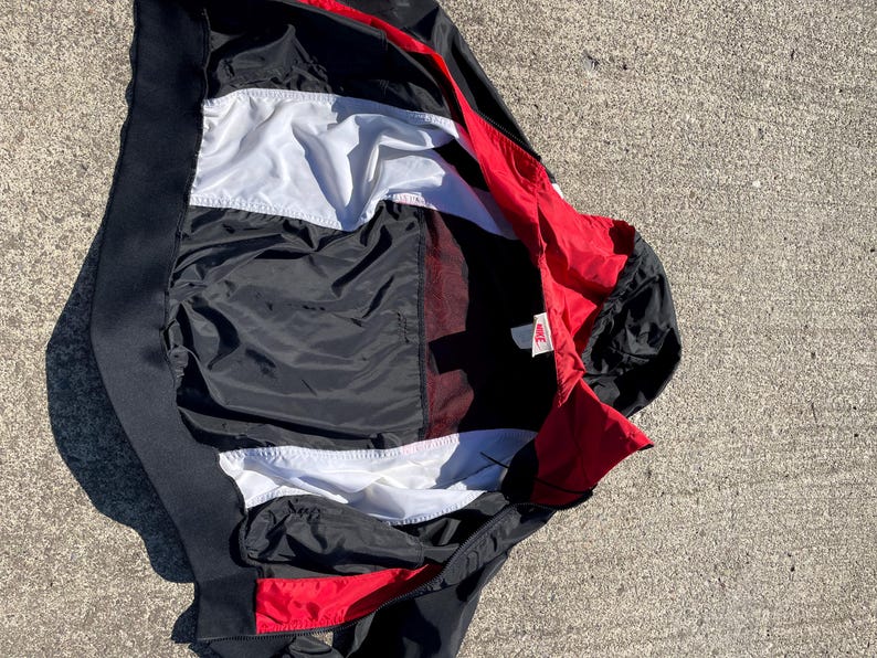 Puede incluir: Una chaqueta cortavientos Nike con un dise&ntilde;o de bloques de color en negro, blanco y rojo. La prenda cuenta con un cuello alto, cierre de cremallera frontal y un forro interior blanco. Una peque&ntilde;a etiqueta de la marca Nike es visible cerca del cuello.