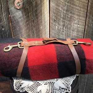 May include: A red and black plaid blanket with a brown leather strap and brass clasps. The blanket is rolled up and ready to be carried.