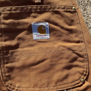 May include: Close-up of brown work pants with a large back pocket. The Carhartt logo is visible on a white and yellow patch. The pants are made of a durable, textured fabric.