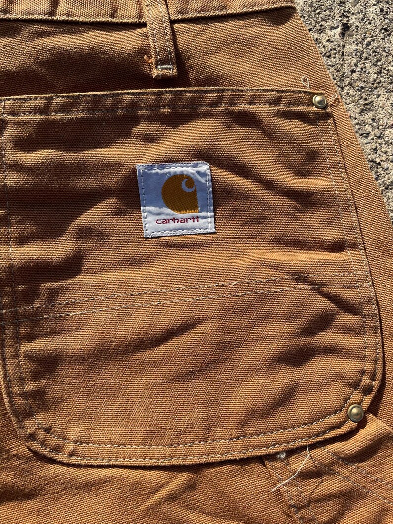 May include: Close-up of brown work pants with a large back pocket. The Carhartt logo is visible on a white and yellow patch. The pants are made of a durable, textured fabric.