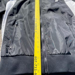 Puede incluir: Chaqueta negra con cremallera y detalles en blanco y gris. La chaqueta tiene un dobladillo y pu&ntilde;os de canal&eacute; negros. Una cinta m&eacute;trica amarilla se coloca verticalmente a lo largo de la chaqueta, mostrando medidas en cent&iacute;metros.