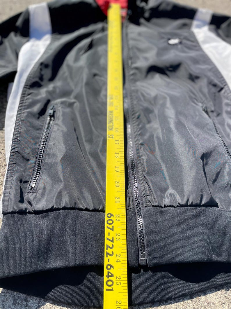 Puede incluir: Chaqueta negra con cremallera y detalles en blanco y gris. La chaqueta tiene un dobladillo y pu&ntilde;os de canal&eacute; negros. Una cinta m&eacute;trica amarilla se coloca verticalmente a lo largo de la chaqueta, mostrando medidas en cent&iacute;metros.