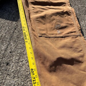 May include: Close-up of brown work pants with a large pocket and a yellow measuring tape. The tape shows measurements in inches, with the phone number 607-722-6401 visible. The pants are made of a durable fabric.