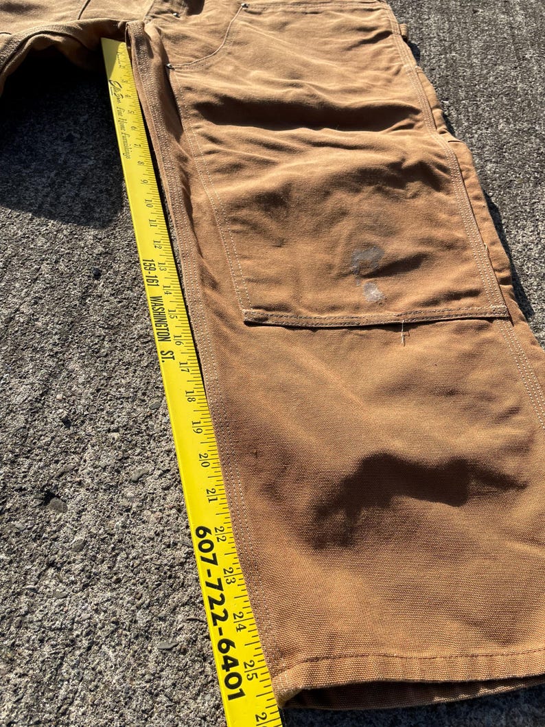 May include: Close-up of brown work pants with a large pocket and a yellow measuring tape. The tape shows measurements in inches, with the phone number 607-722-6401 visible. The pants are made of a durable fabric.