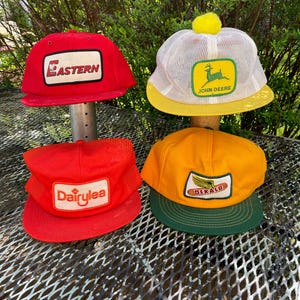 May include: Four vintage baseball caps in red, yellow, and white. The caps feature logos for "Eastern", "John Deere", "Dairylea", and "DeKalb". The John Deere cap has a yellow pom-pom on top.