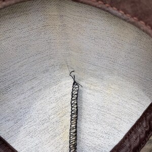 Op de afbeelding: De binnenkant van een bruin stoffen item met een witte, textuurrijke voering. Een enkele verticale naad is verbonden met zwarte overlockstiksels die vanuit het midden naar de bovenrand lopen, waar een bruine stoffen rand de opening omlijst.