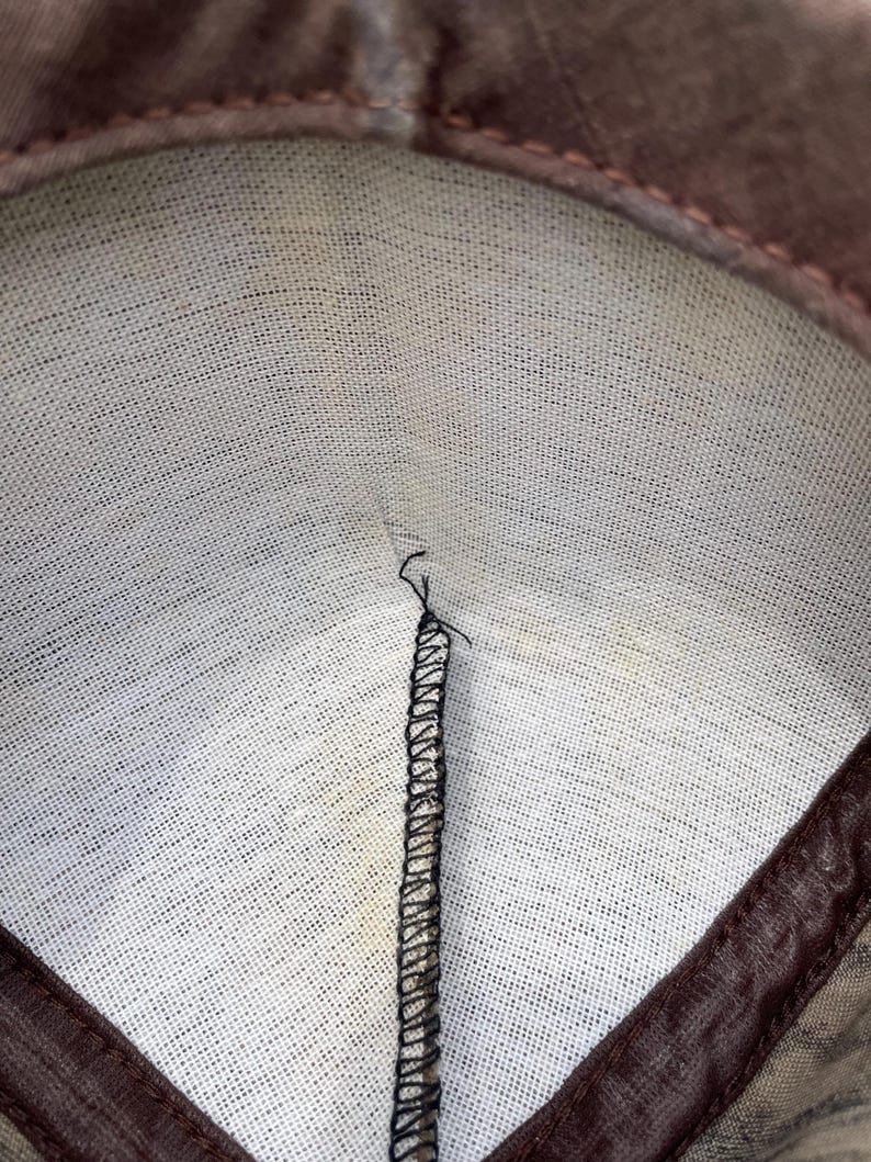 Op de afbeelding: De binnenkant van een bruin stoffen item met een witte, textuurrijke voering. Een enkele verticale naad is verbonden met zwarte overlockstiksels die vanuit het midden naar de bovenrand lopen, waar een bruine stoffen rand de opening omlijst.