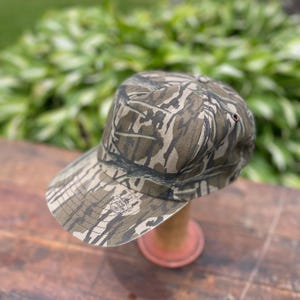 Op de afbeelding: Een baseballpet met een bruin, beige en zwart camouflagepatroon. De pet heeft een gestructureerde kroon en een gebogen klep. Een kleine, ronde logopatch is zichtbaar aan de linkerkant van de klep met de tekst Bottomland erop gedrukt.
