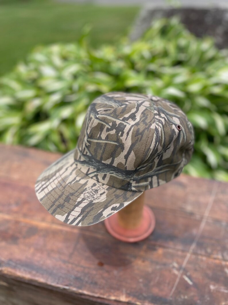 Op de afbeelding: Een baseballpet met een bruin, beige en zwart camouflagepatroon. De pet heeft een gestructureerde kroon en een gebogen klep. Een kleine, ronde logopatch is zichtbaar aan de linkerkant van de klep met de tekst Bottomland erop gedrukt.