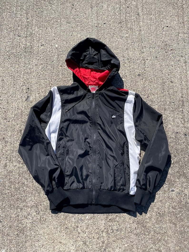 Puede incluir: Chaqueta negra con capucha, forro rojo y rayas blancas laterales. La chaqueta tiene cremallera completa, un peque&ntilde;o logo de Nike y pu&ntilde;os y dobladillo de canal&eacute; negros. Fabricada con un material ligero.