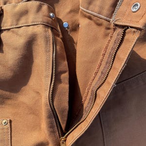 May include: Close-up of brown work pants with a visible zipper and metal rivets. The pants have a classic design with reinforced stitching and a durable fabric, suggesting they are built for heavy-duty use. The zipper is gold-colored.