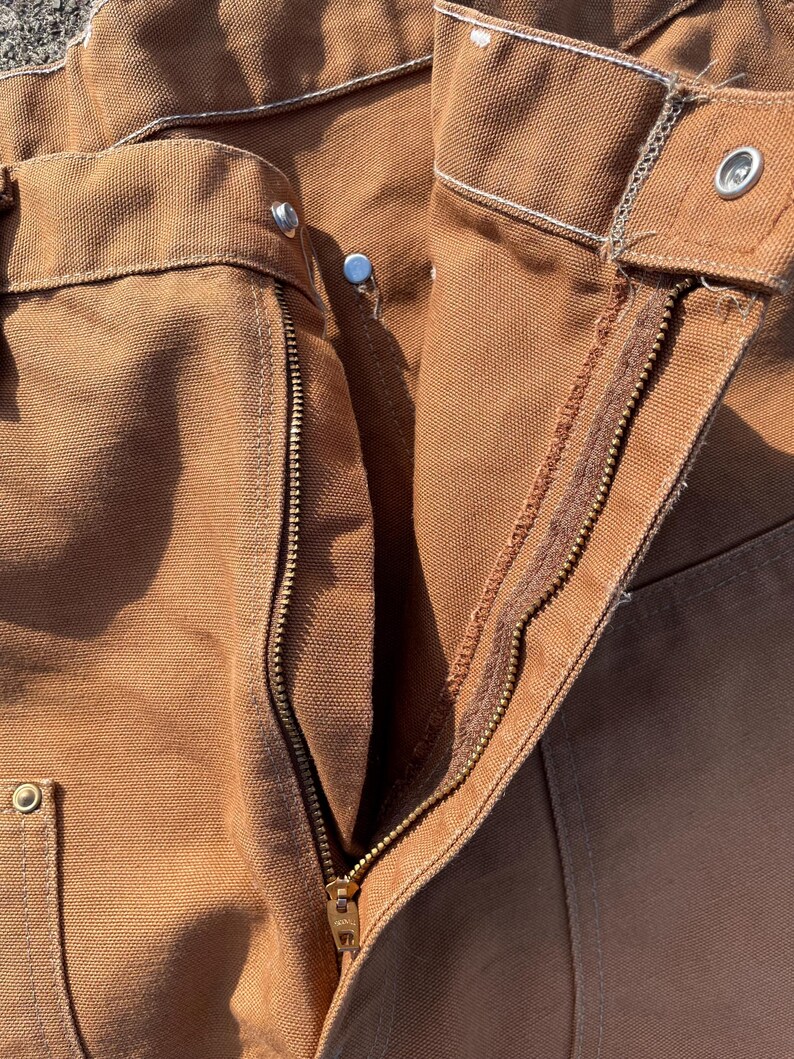 May include: Close-up of brown work pants with a visible zipper and metal rivets. The pants have a classic design with reinforced stitching and a durable fabric, suggesting they are built for heavy-duty use. The zipper is gold-colored.