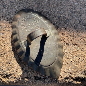 May include: Antique potato peeler with the text "HAMILTON'S POTATO PEELER PAT. PEND'G" embossed on the metal surface. The peeler has a curved handle and a scalloped edge. The metal has a weathered, aged appearance.