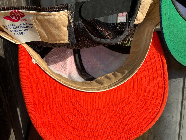 May include: A red baseball cap with a brown mesh back and a white label that reads "100% NYLON - DOUBLE KNIT The "PROFESSIONAL" Baseball Cap LARGE"