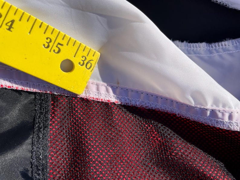 Puede incluir: Una prenda con un panel de tela blanca, una secci&oacute;n negra y una capa de malla roja. Una regla amarilla est&aacute; colocada a lo largo del borde blanco, con la marca de 91,44 cm visible, indicando la medida de la costura del tejido.