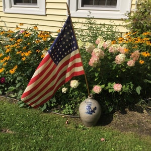 May include: A blue and white ceramic pitcher with a rooster design sits on the ground in front of a flower bed. A small American flag is planted in the ground behind the pitcher.