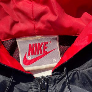 Puede incluir: Primer plano de una chaqueta Nike negra y roja. La chaqueta tiene un forro de capucha rojo y una cremallera negra. El logotipo de Nike se muestra en rojo sobre una etiqueta gris. La chaqueta es talla M.