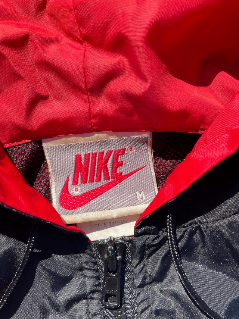 Puede incluir: Primer plano de una chaqueta Nike negra y roja. La chaqueta tiene un forro de capucha rojo y una cremallera negra. El logotipo de Nike se muestra en rojo sobre una etiqueta gris. La chaqueta es talla M.