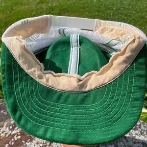 May include: A green and white trucker hat featuring a solid green front panel and brim with a white mesh back. The interior shows a beige sweatband, white mesh lining, and a white plastic snapback closure for adjustable sizing on the back.
