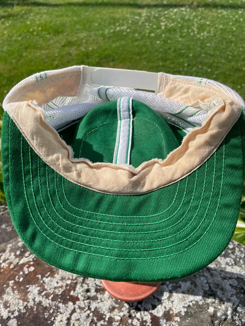 May include: A green and white trucker hat featuring a solid green front panel and brim with a white mesh back. The interior shows a beige sweatband, white mesh lining, and a white plastic snapback closure for adjustable sizing on the back.