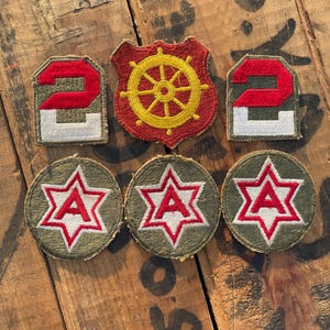 May include: A collection of five embroidered fabric patches. Two rectangular patches feature the red and white number 2 on an olive green background. One shield-shaped patch displays a yellow ship wheel on red. Three circular patches show a red letter A inside a white and red star on olive green.
