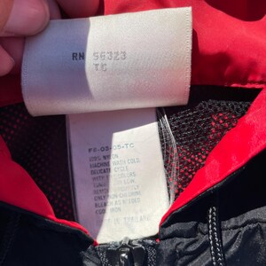 Puede incluir: Primer plano de una chaqueta de nailon negra y roja con cremallera. La etiqueta de la chaqueta muestra el texto "RN 56323 TC" e instrucciones de lavado. La chaqueta est&aacute; hecha de 100% nailon y es lavable a m&aacute;quina.