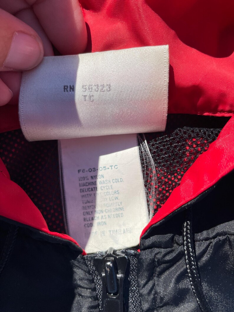 Puede incluir: Primer plano de una chaqueta de nailon negra y roja con cremallera. La etiqueta de la chaqueta muestra el texto "RN 56323 TC" e instrucciones de lavado. La chaqueta est&aacute; hecha de 100% nailon y es lavable a m&aacute;quina.
