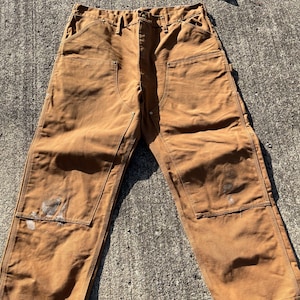 May include: Brown work pants with multiple pockets and reinforced knees. The pants have visible wear and tear, including paint stains. The pants are laid out on a concrete surface.
