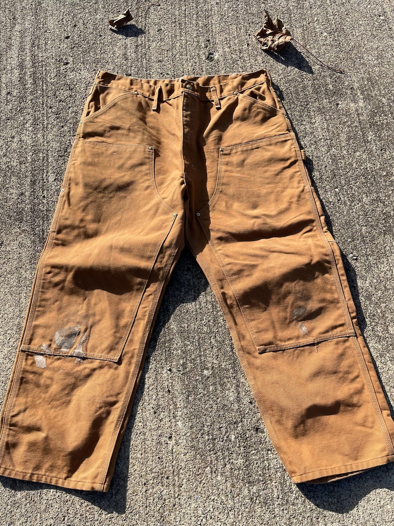 May include: Brown work pants with multiple pockets and reinforced knees. The pants have visible wear and tear, including paint stains. The pants are laid out on a concrete surface.