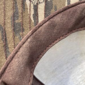 Op de afbeelding: Een close-up van de binnenkant van een baseballpet met een bruine stoffen zweetband en zichtbare stiksels. Het bovenste gedeelte van de pet heeft een camouflagepatroon in bruin-, beige- en donkergroene tinten met de tekst Mossy Oak.
