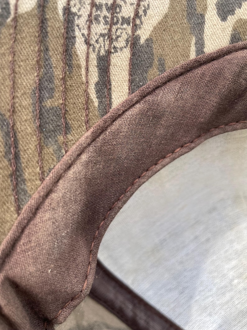 Op de afbeelding: Een close-up van de binnenkant van een baseballpet met een bruine stoffen zweetband en zichtbare stiksels. Het bovenste gedeelte van de pet heeft een camouflagepatroon in bruin-, beige- en donkergroene tinten met de tekst Mossy Oak.