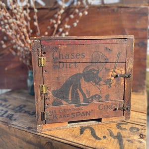 May include: A vintage wooden box with a hinged door and metal latches. The box features a faded graphic and text that reads "Chases Dirt" and "Makes Everything Spick and Span". The box is sitting on a wooden crate.