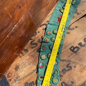 Op de afbeelding: Groene sjerp met meerdere geborduurde patches en badges. Een gele liniaal ligt bovenop de sjerp. Op de sjerp staat het woord "BEA" geborduurd. De sjerp ligt op een houten oppervlak.