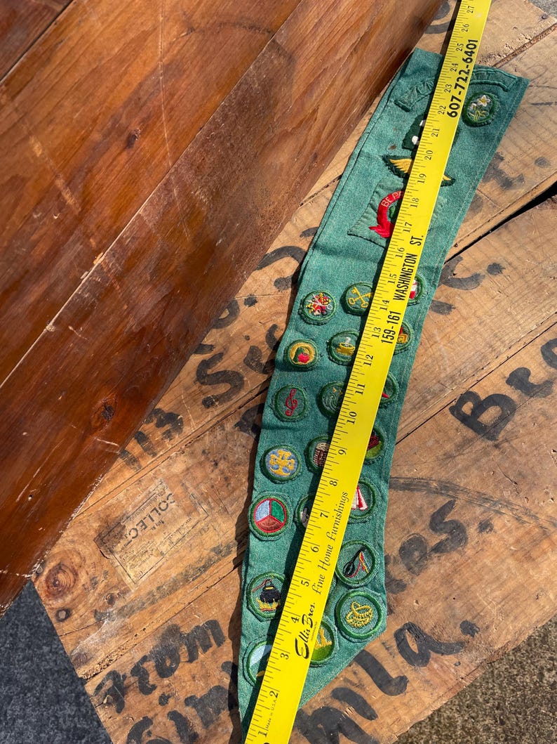 Op de afbeelding: Groene sjerp met meerdere geborduurde patches en badges. Een gele liniaal ligt bovenop de sjerp. Op de sjerp staat het woord "BEA" geborduurd. De sjerp ligt op een houten oppervlak.